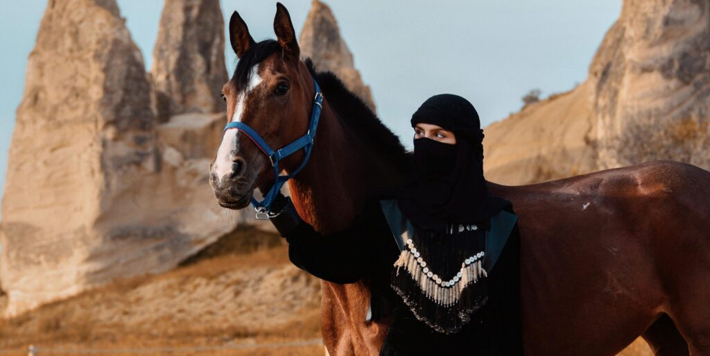 A Middle Eastern woman stands with a horse against a backdrop of unique desert rock formations.