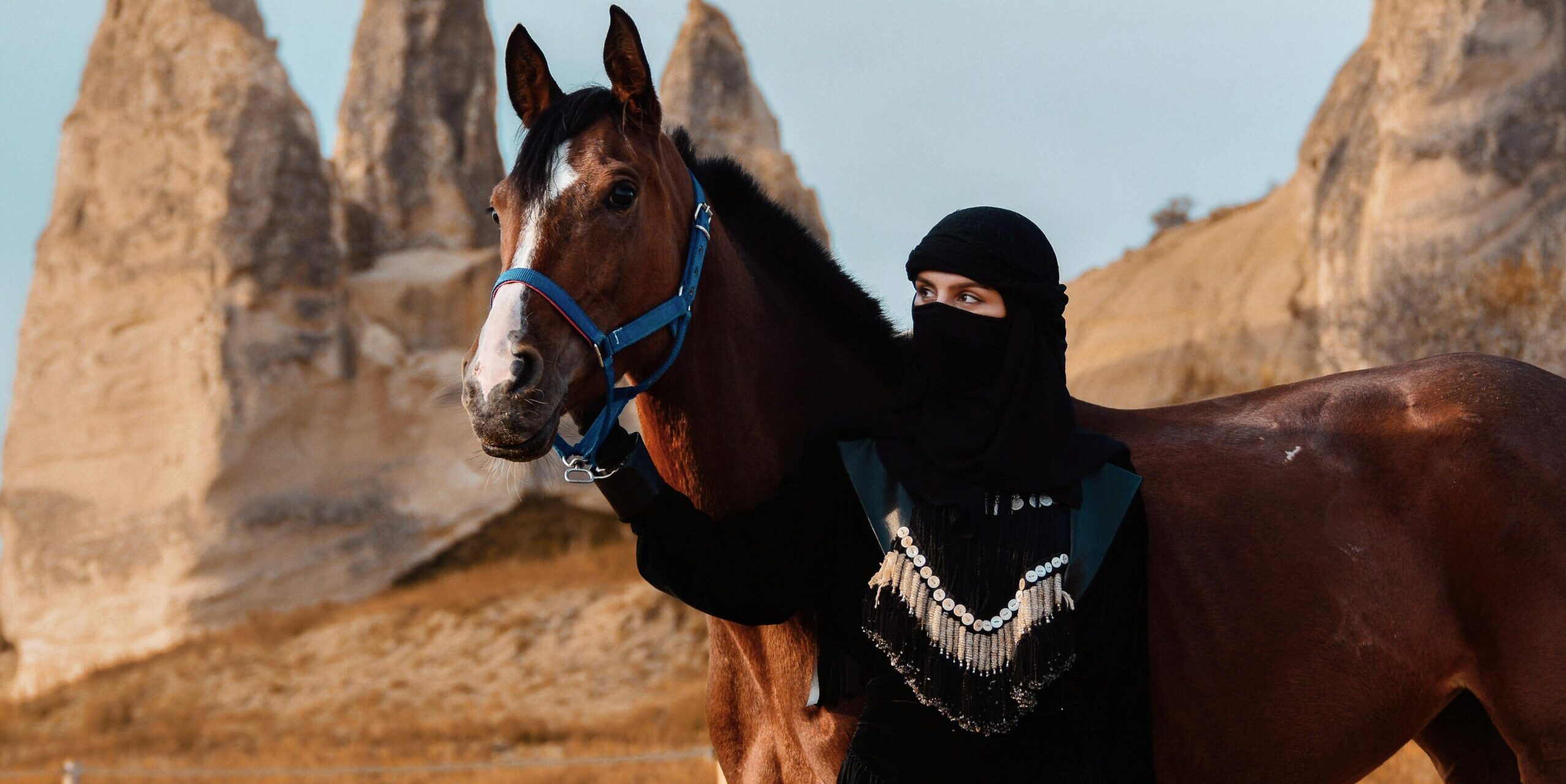 A Middle Eastern woman stands with a horse against a backdrop of unique desert rock formations.