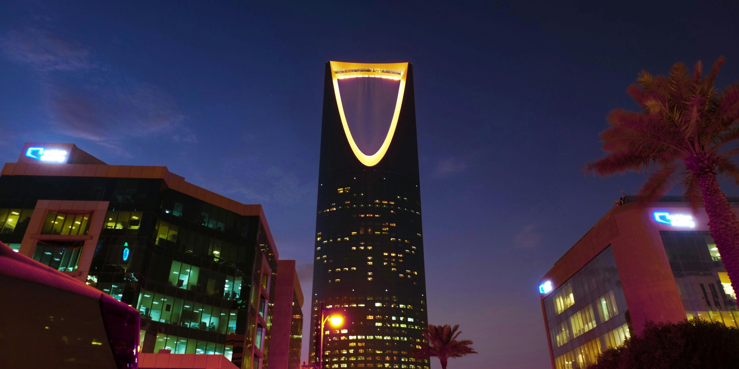 Illuminated Kingdom Centre Tower rises in Riyadh's urban landscape at twilight.
